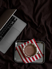 A cup of cocoa on a metal tray, a laptop on brown linens, top view. Lazy morning