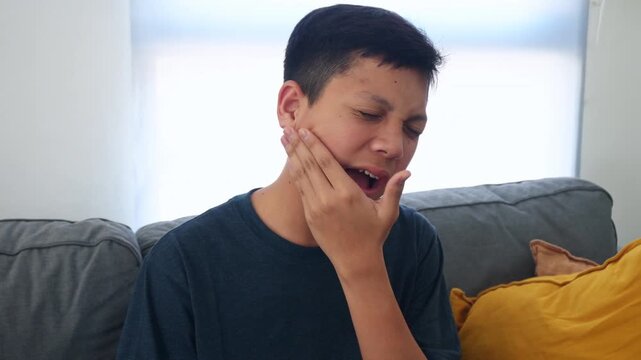 Young Asian Kid Grimaces from Severe Toothache and Clutching Jaw in Discomfort While Sitting on Sofa