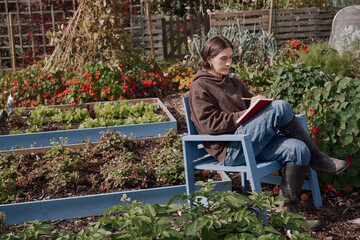 Person writing in red notebook on blue chair in urban allotment garden
