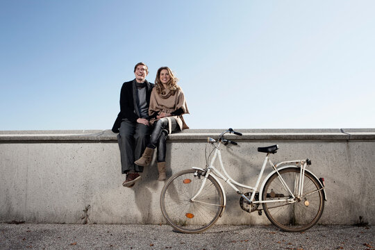 Smiling couple sitting on concrete wall outdoors with bicycle in Munich