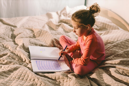 Child sitting on bed drawing in sketchbook at home in pajamas