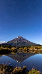 A conical mountain with a snow-capped peak is reflected in a calm, dark lake under a clear starry night sky