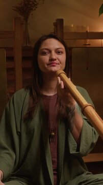 Medium vertical portrait of happy young Caucasian female musician playing didgeridoo flute during esoteric concert or sound meditation session, looking at camera, smiling with tranquility and joy