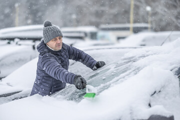 A man in a dark jacket and gray hat diligently removes a thick layer of fresh snow from the windshield of his car with a scraper and a broom. The entire scene takes place during a heavy snowfall 