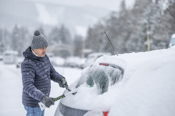 A man in a dark jacket and gray hat diligently removes a thick layer of fresh snow from the windshield of his car with a scraper and a broom. 