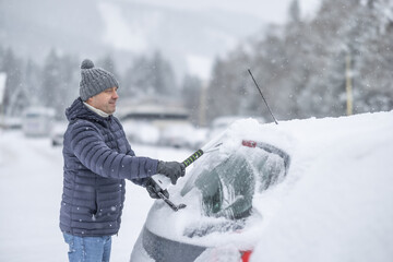 A man in a dark jacket and gray hat diligently removes a thick layer of fresh snow from the windshield of his car with a scraper and a broom. 