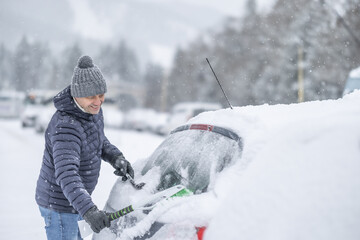 A man in a dark jacket and gray hat diligently removes a thick layer of fresh snow from the windshield of his car with a scraper and a broom. 