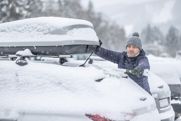 A man in a blue jacket and knit hat uses a broom to clear a thick layer of snow from the back of his car. The surrounding snowy landscape and the roof box on the vehicle suggest that 