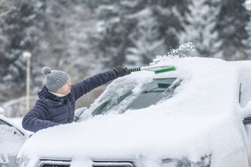 A man in a dark jacket and gray hat diligently removes a thick layer of fresh snow from the windshield of his car with a scraper and a broom. 