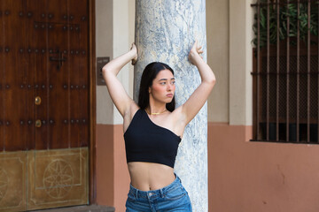 A serious, young, attractive woman with dark hair, dressed in jeans and a black top, leaning back and holding onto the grey marble column in the centre of Seville with her hands.