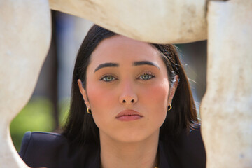 Close-up portrait of a young, beautiful woman with dark hair and blue eyes through a gap in a white marble lion sculpture in a park square.
