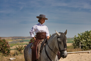 A young, beautiful Spanish woman dressed in riding clothes and wearing a traditional Spanish hat, riding a horse in the countryside on a sunny day.