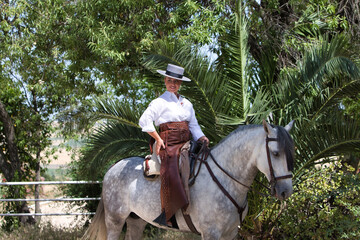 A young, attractive Spanish woman dressed in riding attire and wearing a traditional Spanish hat, riding a horse in the countryside on a sunny day. In the background, green vegetation.