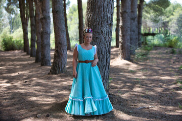 A beautiful young Spanish woman dressed in a traditional green flamenco dress and with a flower in her hair walks through the pine forests of Andalusia in the pilgrimage.