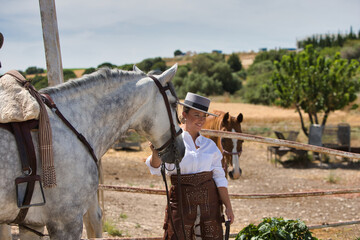 A young, beautiful Spanish woman dressed in riding clothes and wearing a traditional Spanish hat stands next to a horse in the countryside on a sunny day. More horses graze in the background.