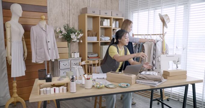 asian female fashion designers walking in modern clothing studio checking dresses on rack discussing ideas surrounded sewing tools fabrics packaging startup workspace creating handmade apparel