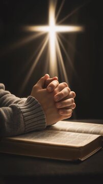 Vertical video of hands clasped in prayer over an open Bible. Glowing cross with divine light rays in the background. Christian faith, worship, and spiritual devotion concept