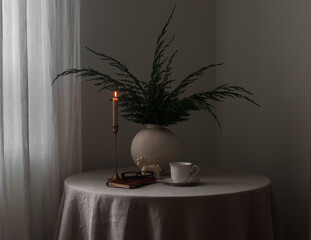 A round table with a gray tablecloth, a vase with juniper branches, a candle, and a cup of coffee in the living room