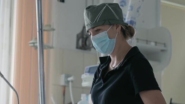 Portrait of focused female doctor or nurse wearing protective medical mask and surgical cap working with patient in hospital operating room or intensive care unit