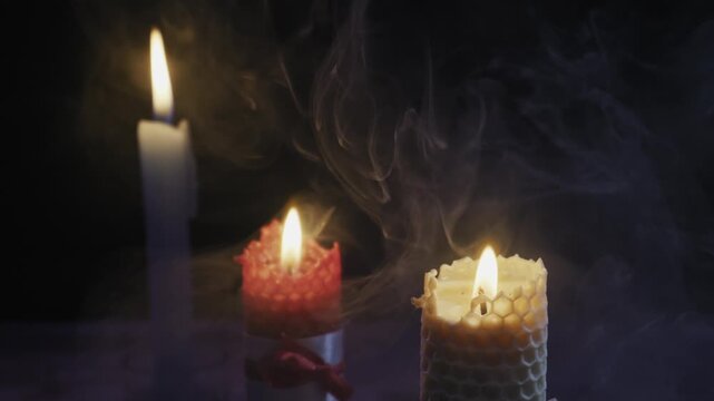 Three lit candles burn against a dark background as mysterious smoke swirls around them. Concept of magic, spirituality, meditation, remembrance, and occult rituals.