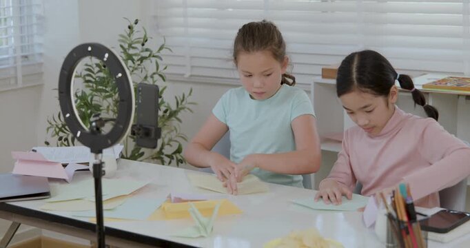 Two diverse kid vloggers teach origami live, folding paper for a DIY tutorial with a phone and ring light at home. This 4k footage is a cinematic clip about e-learning and creativity.