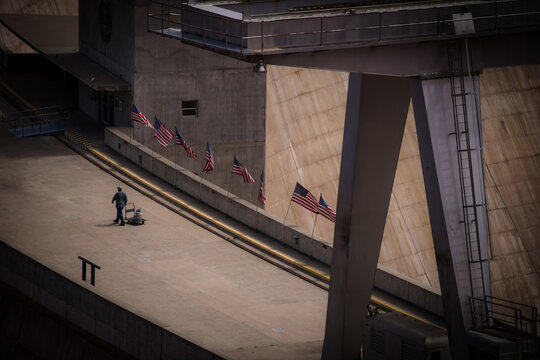 View of a lone worker polishing a concrete walkway adorned with American flags, juxtaposed against the towering Glen Canyon Dam, Page, Arizona, United States.