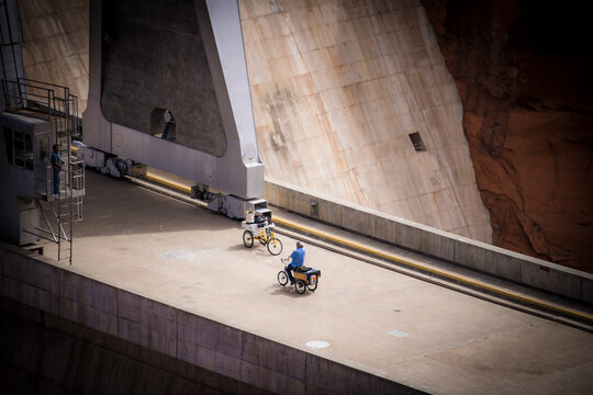 View of cyclists traverse the concrete expanse of the Glen Canyon Dam, its massive scale dwarfing the figures against the red rock backdrop, Page, Arizona, United States.