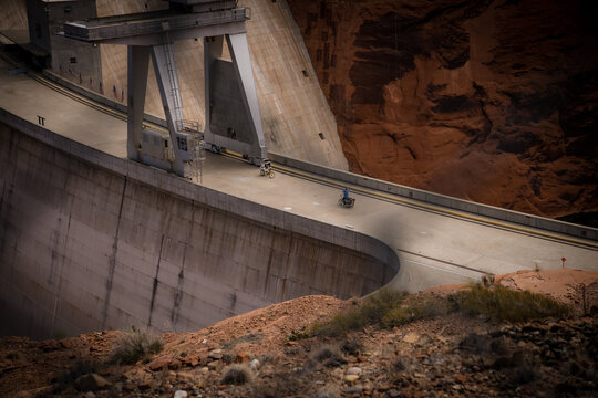 View of the immense concrete Glen Canyon Dam curves against the stark red rock landscape, with figures crossing, a testament to human engineering, Page, Arizona, United States.