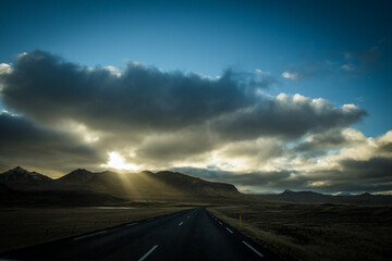 View of the road stretches into the distance, the sun's rays breaking through dramatic clouds over rugged mountains, creating a serene landscape, Grundarfjordur, West Iceland, Iceland.