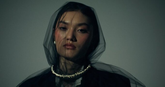 A serious fashionable woman looks at the camera posing in the studio, close-up of her face. A black veil on her face. The light turns on and off