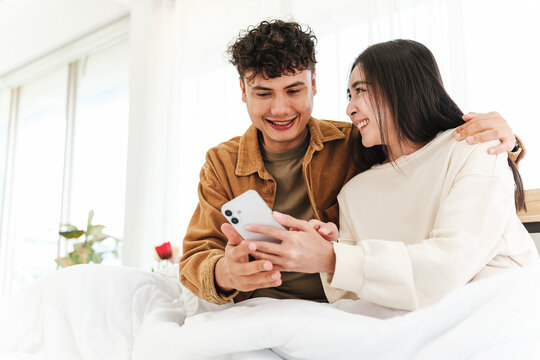 Young Asian couple sits closely on a cozy bed, smiling and looking at a smartphone together, capturing warmth, digital connection, relaxation, and affection in a bright modern bedroom. - Powered by Adobe
