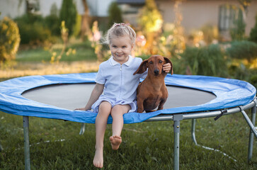 a three year old smiling girl sitting with dachshund dog on trampoline in green sunny summer park