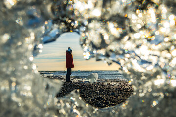 View of a person in a red coat stands on black sand beach framed by sparkling ice formations in Jokulsarlon Diamond Beach, Hofn, Iceland.