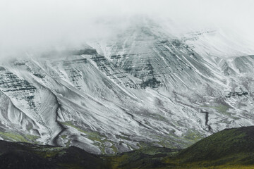 View of a snow-dusted mountain with rugged, dark rock formations and patches of green vegetation under a misty sky, Blonduos, Vatnsnes peninsula, Iceland.