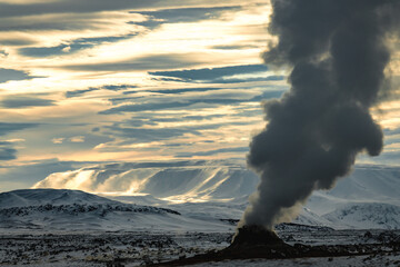 View of a geothermal area's plume of steam rising against a backdrop of snow-covered hills under a dramatic sky, Rejkjahlid, North Iceland, Iceland.