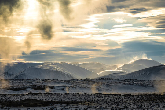 View of the Namafjall Geothermal Area's stark, snow-dusted landscape where ethereal steam rises against a backdrop of undulating hills under a radiant sky, Rejkjahlid, Iceland.