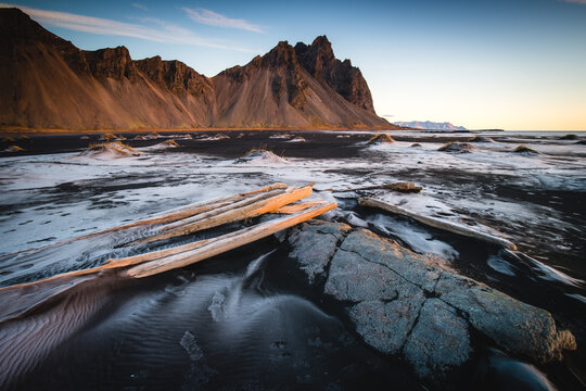 View of jagged mountains rising from a black sand beach with driftwood and textured patterns carved by the tide, Hofn, Iceland.