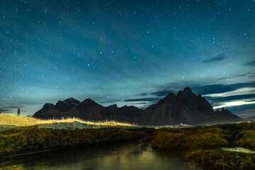 View of dark, jagged mountains silhouetted against a starry sky with wispy clouds over a reflective stream and golden grasses, Hofn, Iceland.