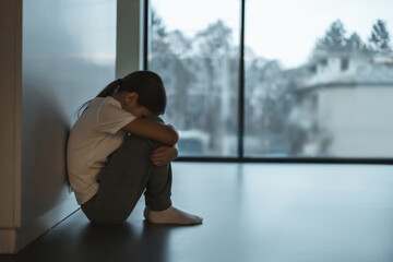 Lonely child huddled in corner by large window expressing kid depression and fear of bullying abuse