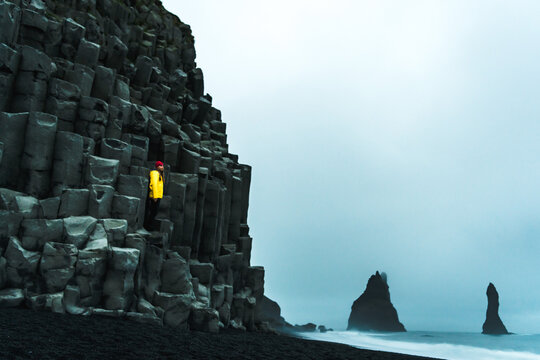 View of a solitary figure in a bright yellow jacket perched on basalt columns, contrasting against the dark sand and misty sea stacks, Vik, Iceland.