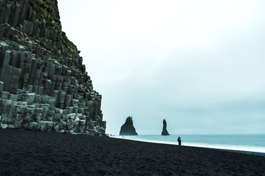 View of basalt columns rising sharply from the black sands as a lone figure gazes towards the sea stacks in the distance, Vik, Iceland.