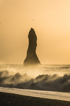 View of basalt sea stacks stand tall against the crashing waves in a mystic sea, where the golden light paints the scene in Reynisfjara, Vik, Iceland.