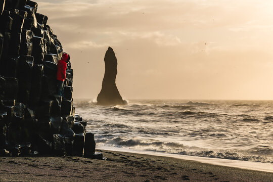 View of a person in red jacket climbing black basalt columns next to the roaring sea with a monolithic sea stack rising from the waves, Vik, Iceland.