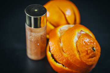 A partially peeled orange next to a shiny cosmetic bottle with a metallic cap on a black...