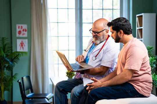 Indian male doctor using laptop while consulting young patient during clinic visit