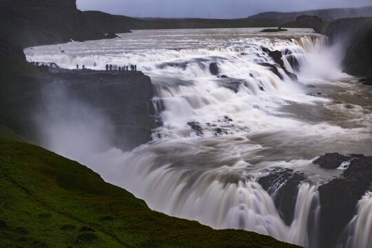 View of the powerful Gullfoss waterfall cascading over rugged cliffs into the canyon below, with tourists enjoying the vista, Haukadalur valley, Iceland.