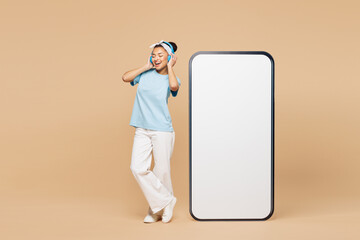 Full body young smiling happy woman wear blue t-shirt casual clothes stand big huge blank screen mobile cell phone smartphone with area listen music in headphones isolated on plain beige background.