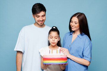 Young happy parents mom dad with child kid daughter girl wear casual clothes hold in hand cake puff out candles making wish isolated on plain blue background. Family day parenthood childhood concept.