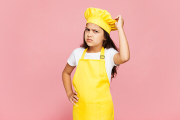 Small confused puzzled minded kid child girl housewife chef cook baker mother's helper wear yellow apron toque cap scratch head temple isolated on pastel plain pink background. Cooking food concept.