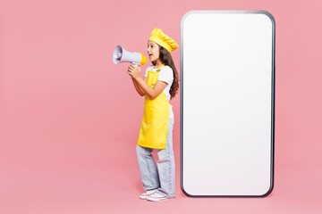 Full body small kid child girl housewife chef cook baker mother helper in yellow apron toque cap big blank screen area mobile cell phone scream isolated on plain pink background Cooking food concept